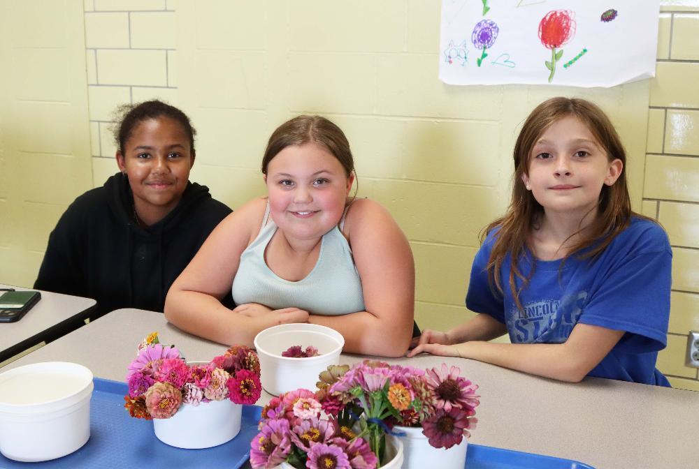 Three Dawes Middle School students are sitting at a table in the school cafeteria. The three girls are sitting behind bouquets of zinnias that are in circular pots. Three of the pots are full of flowers and a fourth one is halfway full of zinnias. The students were helping with Family Engagement Night activities at school during the evening.