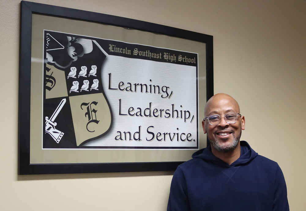 Eric Williams smiles next to a framed poster in a Southeast High School office. The poster says, 'Lincoln Southeast High School: Learning, Leadership and Service' and has a picture of the school coat of arms.