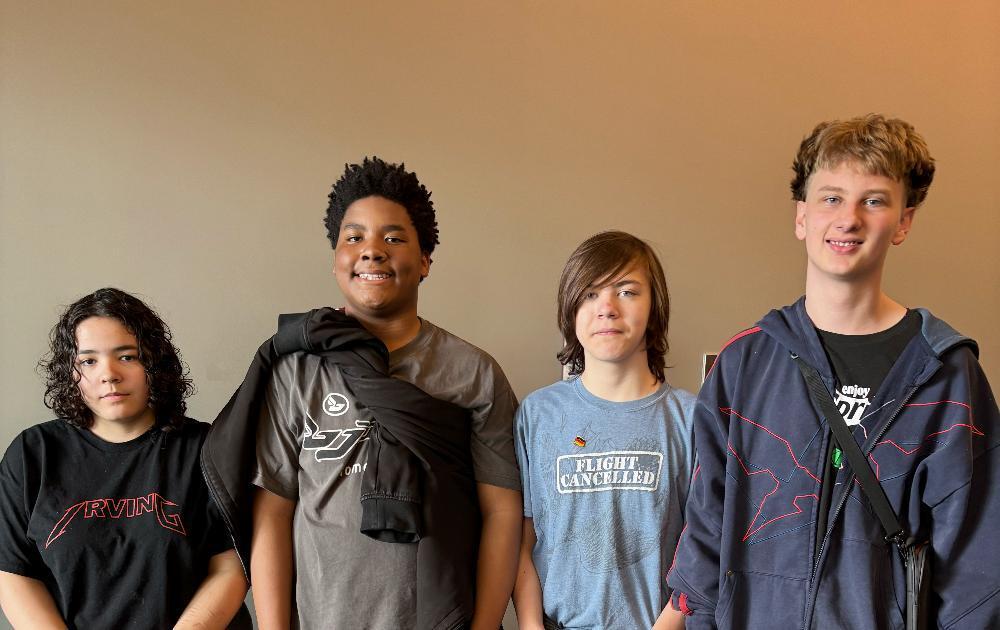 Four diverse young people stand side-by-side against a plain wall. Two smile, while others have neutral expressions. They wear casual t-shirts and hoodies, including one with 'Flight Cancelled' and another with a jacket draped.
