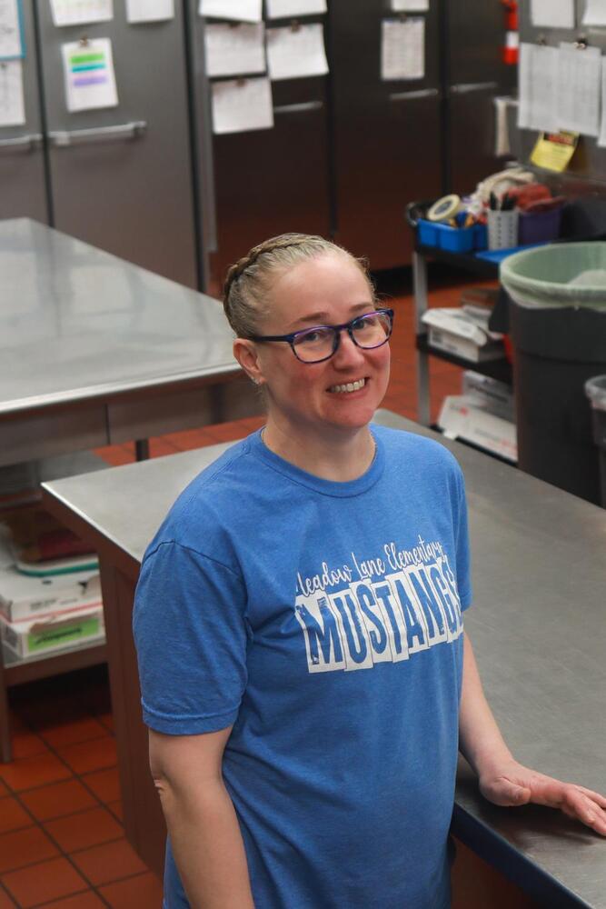 A smiling woman in a blue "Meadow Lane Elementary Mustangs" t-shirt stands in a school kitchen. She has glasses and braided hair, with her hand on a stainless steel counter. Stainless steel refrigerators and tables are in the background.