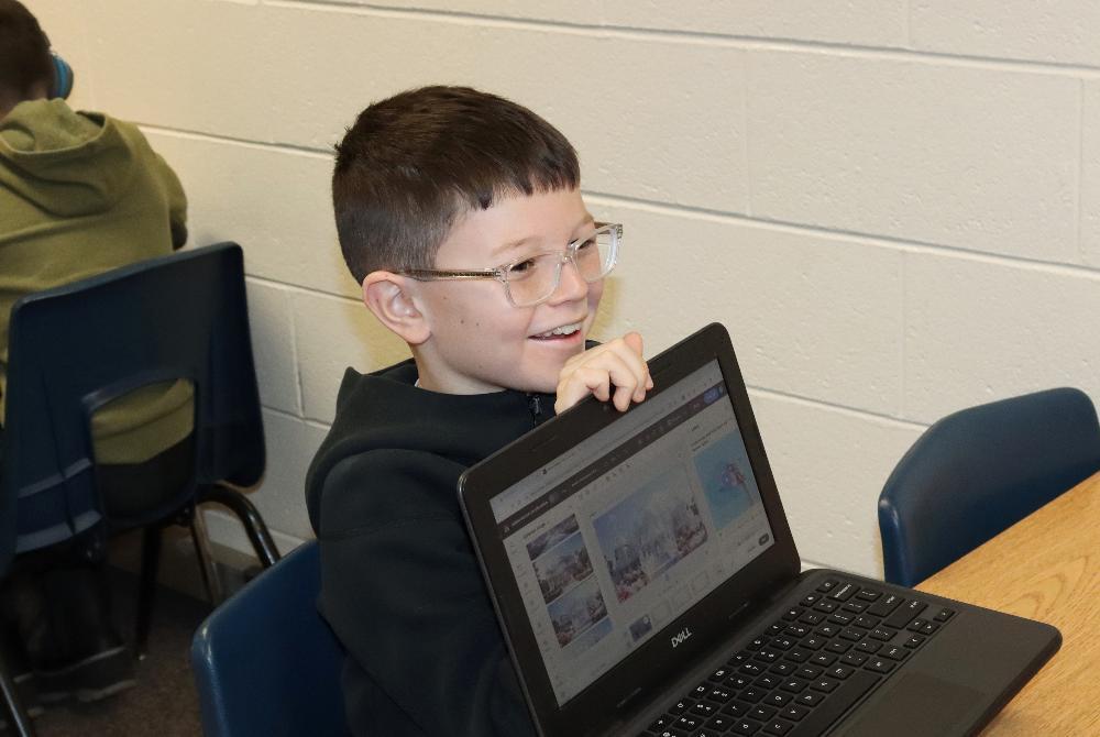 A Cavett Elementary School fifth grader smiles as he displays a project on a computer screen. He is wearing glasses and has turned his laptop around to show an image created by generative artificial intelligence. The image is of a castle covered in snow during winter. The Cardinals worked with the generative AI program during a computer science class.
