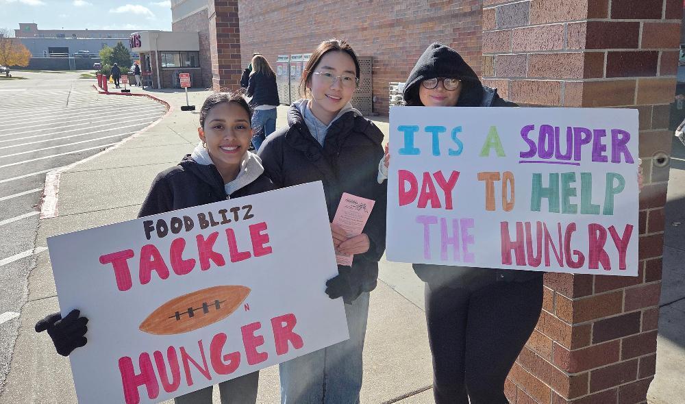 Three Southwest High School students are standing in a row outside a local grocery store for the Food Blitz XIII event. A girl on the left side of the row is holding a sign that says Food Blitz Tackle for Hunger. The girl in the middle is holding a pink delivery card, and the girl on the right is holding a sign that say It's a Souper Day to Help the Hungry.