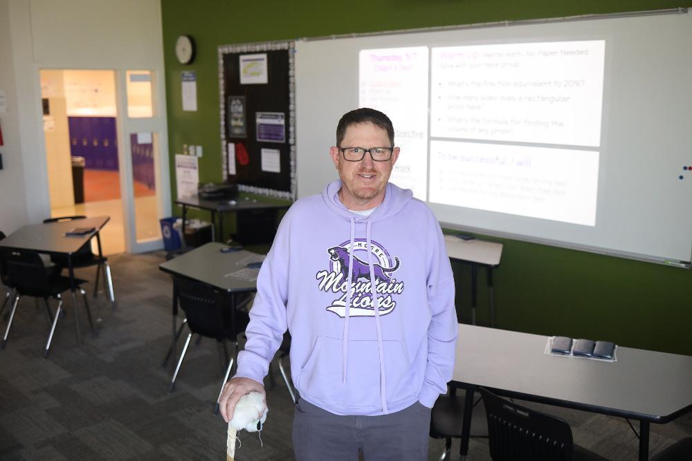 A smiling man in glasses and a purple "Mountain Lions" hoodie stands in a classroom, holding a white bird on his left hand. Desks, chairs, a whiteboard with math problems, and a hallway with purple lockers are visible in the background.