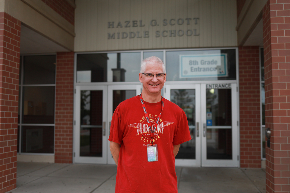 Man with white hair and glasses standing in front of a  school smiling.