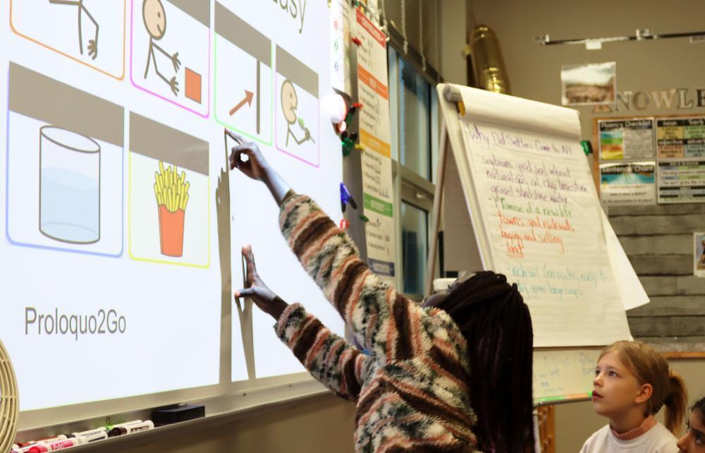 A fourth grade student at Huntington Elementary School uses her left hand to point to a symbol on a screen in the classroom. She and other students are trying to decode a row of symbols that are on an augmentative and alternative communication device. There are four symbols on the top row and two on the bottom row that stand for a sentence of "I want to eat some french fries."