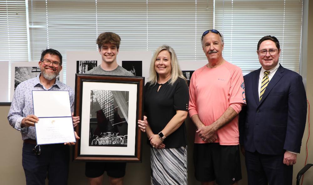 A framed black and white photograph of bicycles in an architectural setting is being presented. A young man receives an award certificate from a man, with family and a man in a suit observing, in an office setting.
