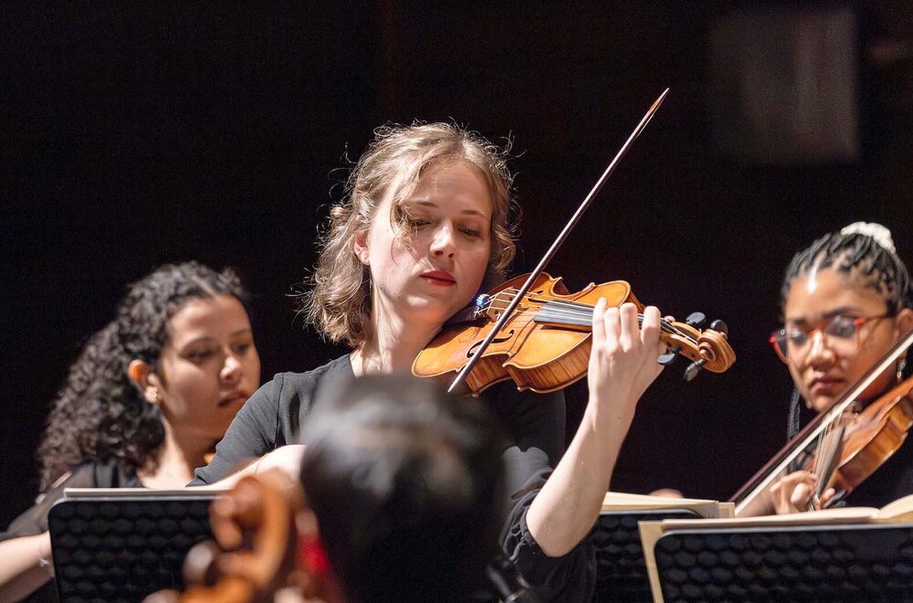 Sheridan Elementary School graduate Annemarie Schubert plays her violin during an orchestra practice. She is standing in the middle of two other players. She is holding her violin in her left hand and the bow in her right hand.