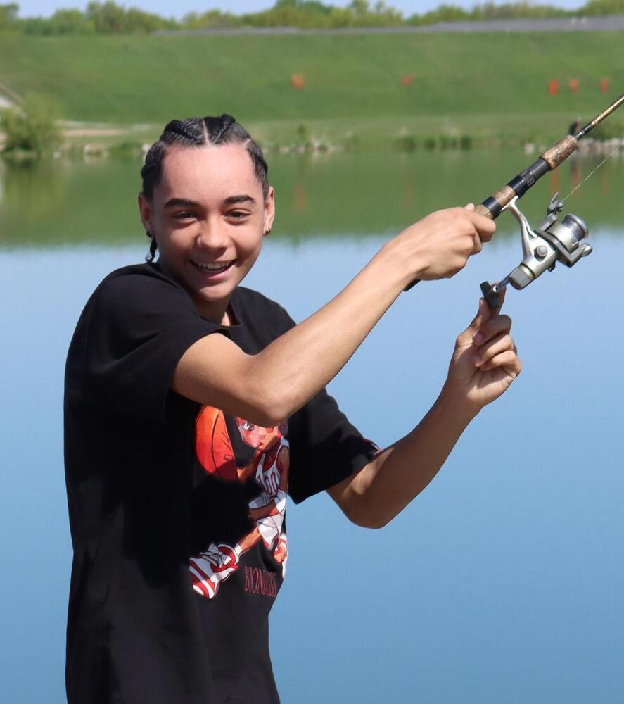 A young person with braided hair holds a fishing rod near a calm lake. They are smiling.