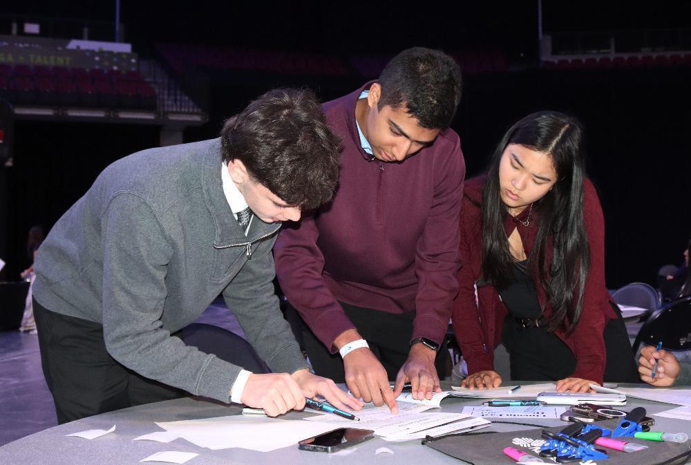 Three students, two young men and one young woman, lean over a table. They are intently focused on papers, pointing and writing with pens, seemingly collaborating on a task at an event.