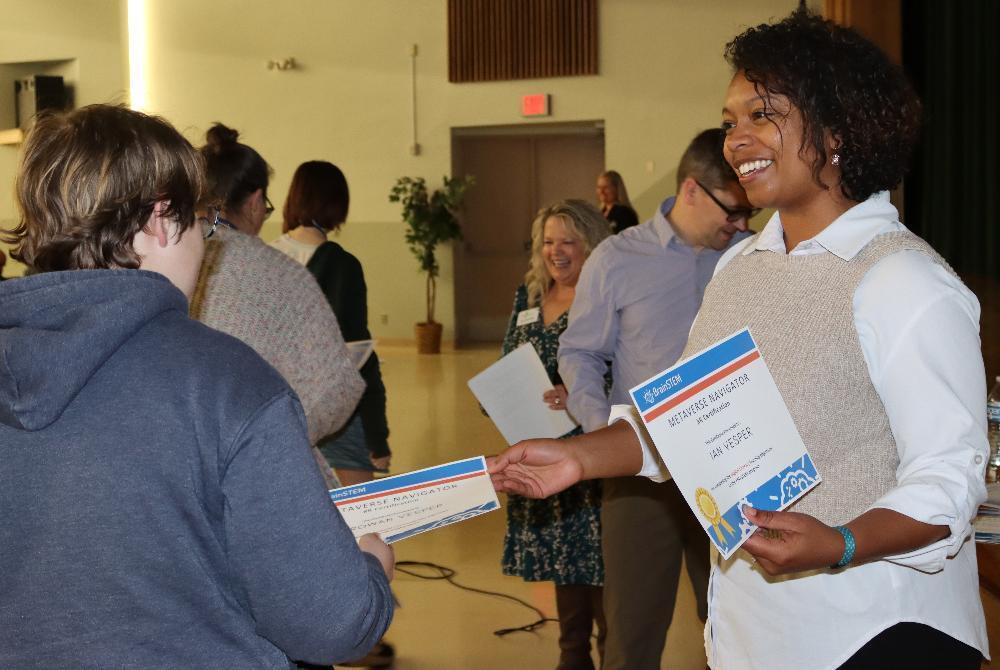 A smiling woman hands a "Metaverse Navigator" VR certificate to a person in a hoodie, their back to the camera. Several people in the background are smiling, suggesting a lively certification ceremony.