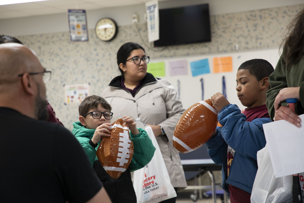 Two students hold inflatable footballs at a CLC signup event. Two adults are standing next to them in a school cafeteria.