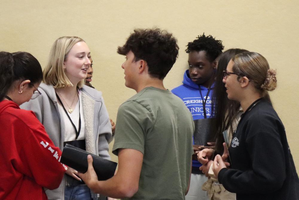A group of teenagers converse; a blonde girl holds a black wallet. They stand in a beige-walled room. One person's sweatshirt says "CREIGHTON JUNIOR HIGH."