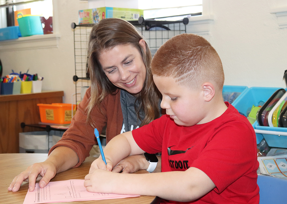 Norwood Park paraeducator Gemma Love smiles as she works with a student at a classroom table. The student is using a pencil to write on a paper worksheet laying on the table. Gemma is sitting next to him and is looking at the worksheet.