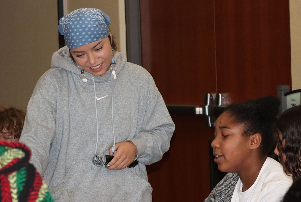 From left, national music composer and producer Olivia Komahcheet smiles as she talks with a middle school student in the LPS district office boardroom. Olivia is standing next to a table and is holding a microphone in her left hand. The student is talking to her at the table.