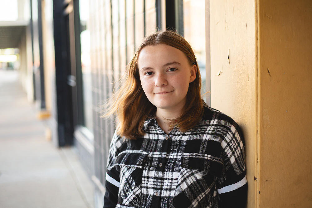 A teenage girl, wearing a black and white plaid shirt, stands leaning against a building's exterior wall.