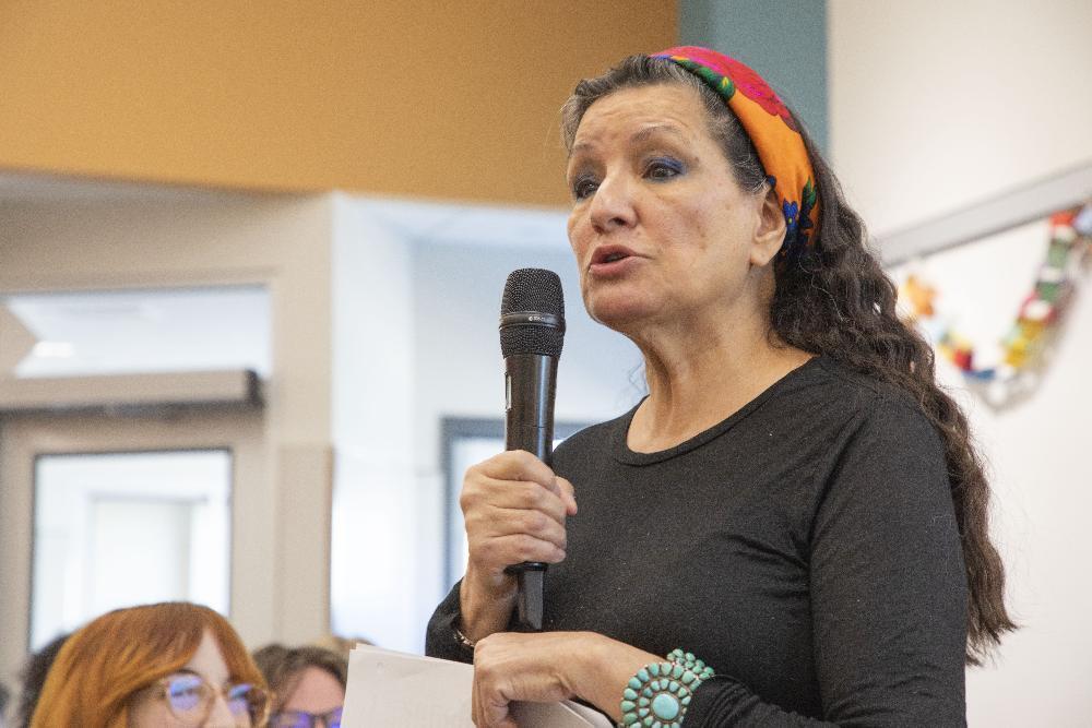 A woman with long dark hair and a colorful headband speaks into a microphone, wearing a black shirt and turquoise bracelet. Her mouth is open mid-sentence at an indoor event.