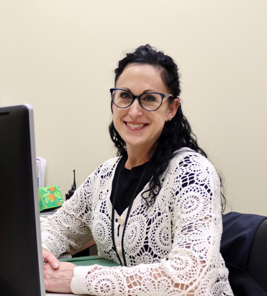 LPS Security Coordinator Sarah Balcom smiles in her office at the Steve Joel District Leadership Center. She is sitting at her desk in the center of the photo, and a computer screen is on the far lefthand side of the photo. She is wearing glasses and a long-sleeved outfit.