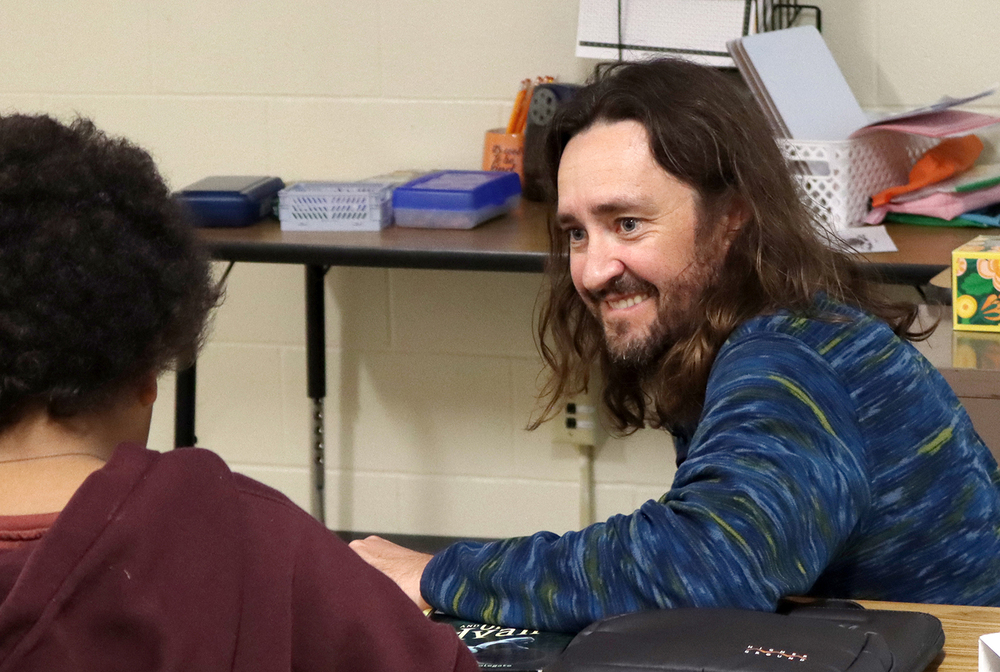 Park Middle School teacher Tanner Hilzer smiles while talking with a student in his classroom. He is crouched down next to the desk to reach the same eye level as the male student.