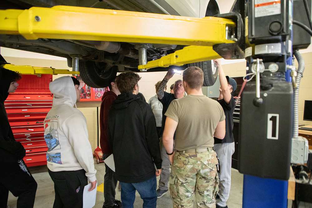 Northeast High School teacher Andy Christensen shines a flashlight at the underbelly of a car while seven students stand with him under the raised car. The car is raised above their heads on an auto mechanic lift system. They are studying how car engines work.