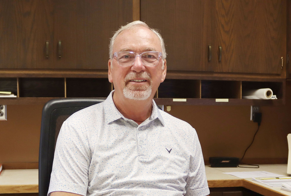 LPS Supervisor of Maintenance Tim Loseke smiles while sitting in a chair in his office. There is a row of cubbyholes hanging below two doors for shelving on the wall behind him.