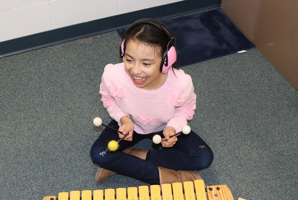 A Riley Elementary School student beams as she plays the xylophone in class. She is sitting down next to the instrument and is holding mallets in both hands.