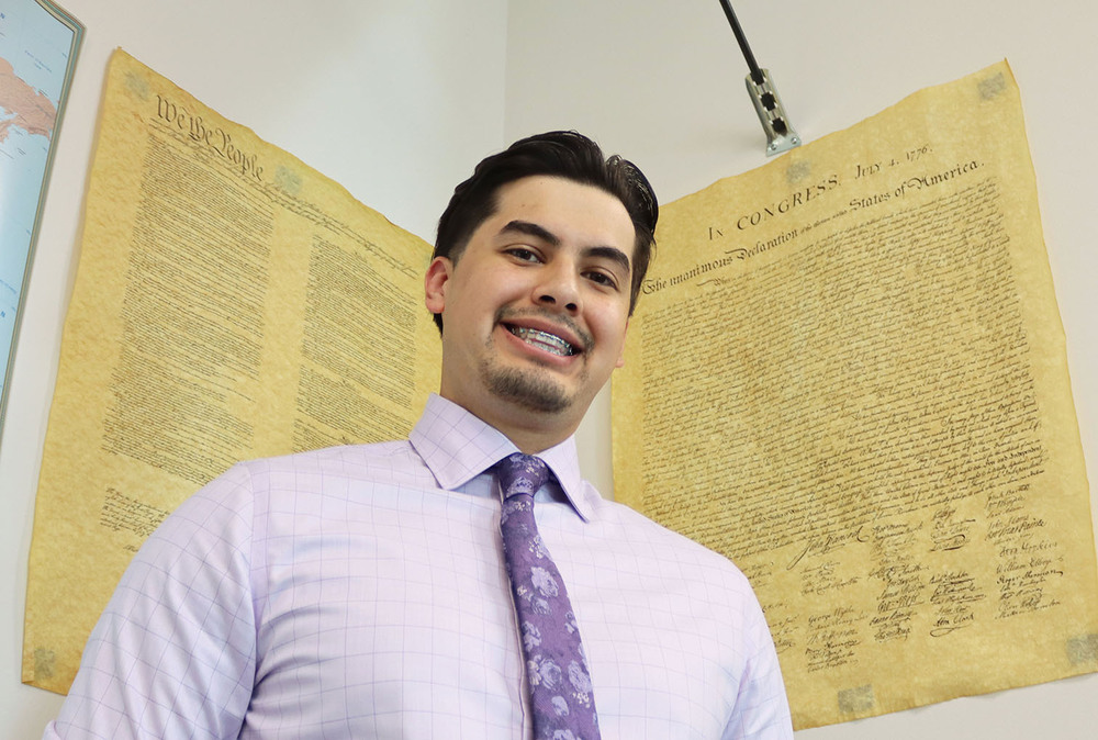 Northwest social studies teacher Edgar Torres smiles in front of two posters that serve as copies of the United States Constitution. The posters are on two corners of a wall in his classroom. He is wearing a shirt and tie that have Northwest's school colors on them.