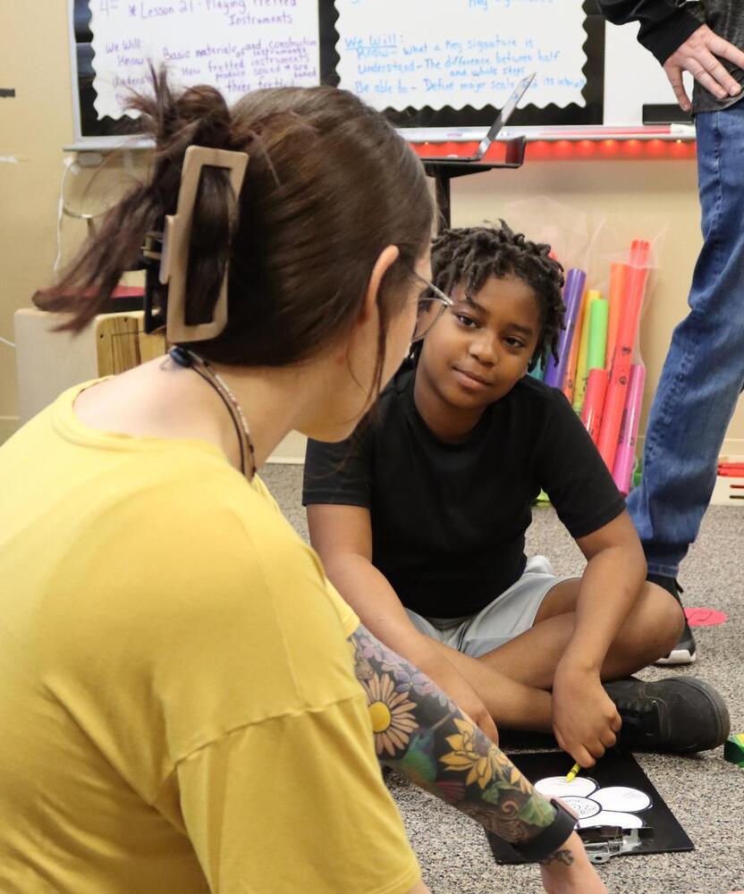 A woman with a yellow shirt and a floral tattoo assists a boy; he sits cross-legged on the floor completing a worksheet. The context is a classroom setting.