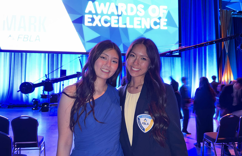 Two East High School students are smiling at the FBLA National Leadership Conference. Both girls are standing in front of a large screen that says Awards of Excellence on it.