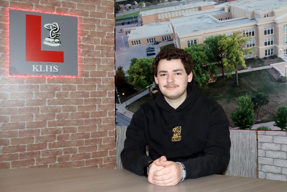 A curly-haired young man with a mustache, wearing a black hoodie, sits at a table. Behind him, a brick wall features a red-lit "KLHS" sign next to a large mural showing an aerial view of a school campus.