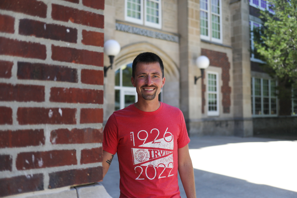 Smiling man in. a red shirt standing in front of a brick school building.