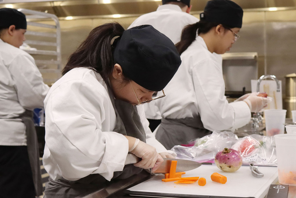 A North Star student uses a kitchen knife to cut up pieces of carrots during a state competition. A second student is washing a vegetable behind her and two other students are working behind them.