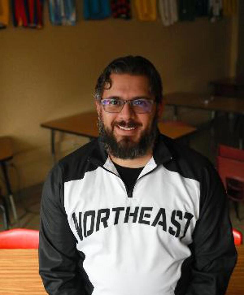 Smiling man with glasses and a beard wearing a Northeast pullover, seated in a classroom.