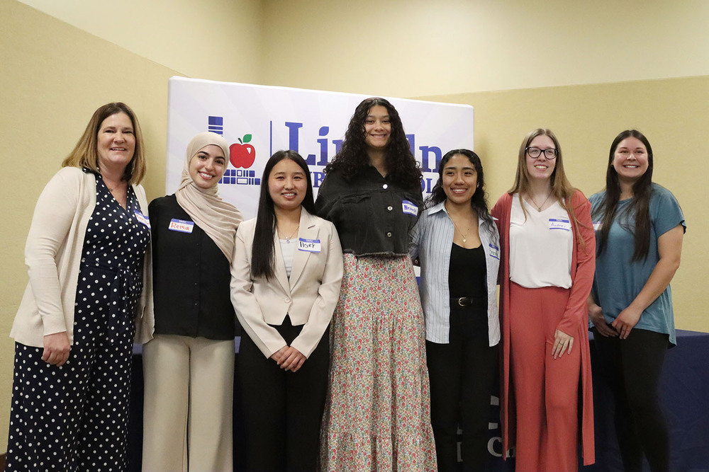 One teacher and six future early childhood teachers smile together while standing in a row in the district office boardroom. They are standing in front of a banner with the LPS logo on it.