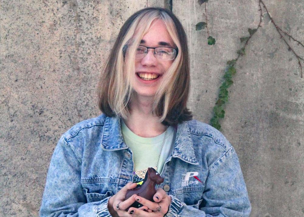 A person with shoulder-length blonde hair, wearing glasses and a denim jacket, holds a small brown horse figurine. The backdrop is a textured concrete wall with a vine.