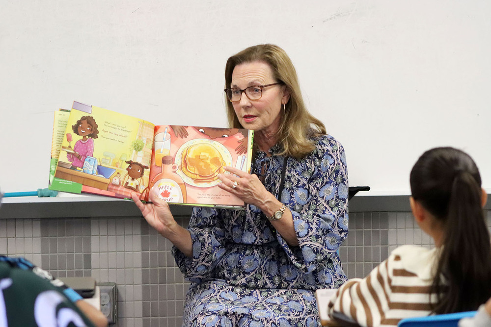 Nebraska First Lady Suzanne Pillen displays two pages of a book to a class of McPhee Elementary School fifth graders. the book pages show a mother and daughter preparing to eat a stack of pancakes with maple syrup.