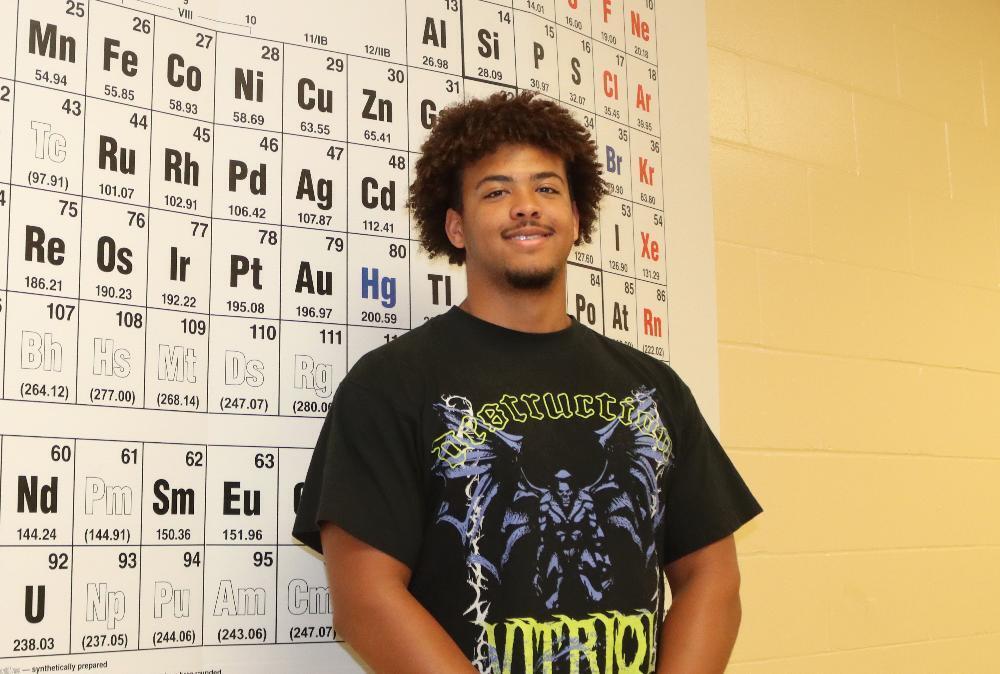 A smiling young man with dark curly hair, wearing a black graphic t-shirt, stands in front of a large periodic table. The background suggests a science classroom.