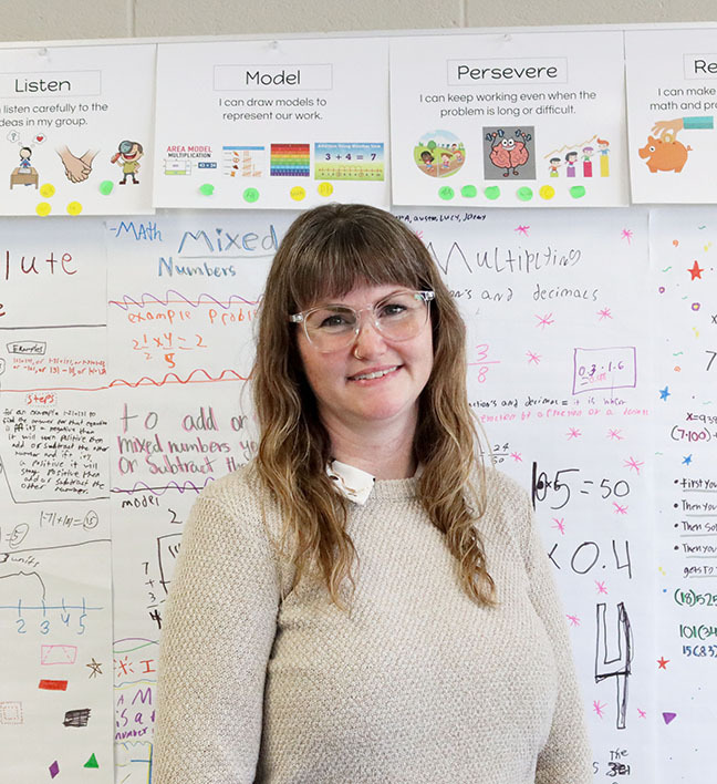 Dawes teacher Amy Kinnett smiles in front of a wall that contains math posters done by students. There are three signs near the ceiling with the words Listen Model Persevere on them.