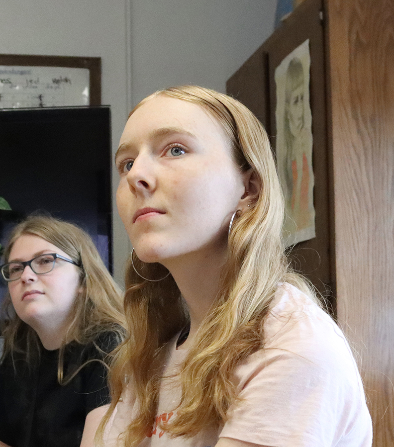 Phoebe Pierson listens to a presentation in her world language class. A classmate is sitting behind her at a desk. Both are facing right to left.