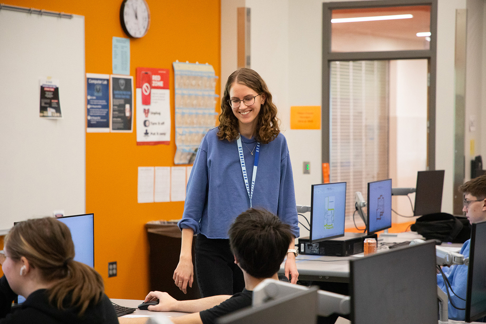 Alex Flamme smiles as she speaks with Standing Bear High School students. The students are sitting in front of a row of computers in a classroom.