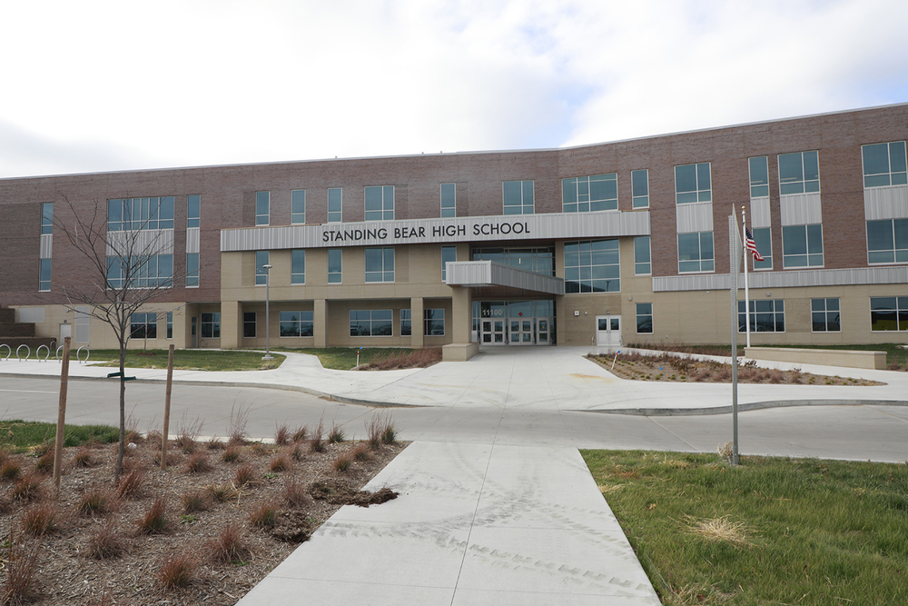 The photo shows the exterior of Standing Bear High School. The three-story building is behind a bus lane and sidewalk. The words Standing Bear High School are above the entryway.