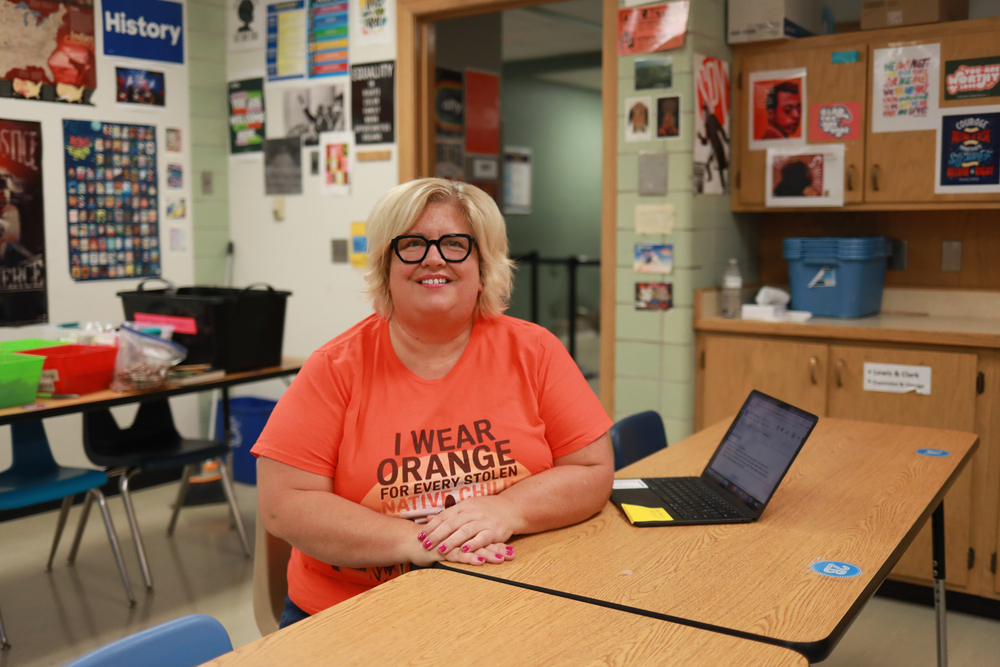 A woman wearing glasses and an orange shirt sits at a classroom table beside an open laptop, smiling at the camera. Posters and student materials decorate the classroom walls and cabinets.