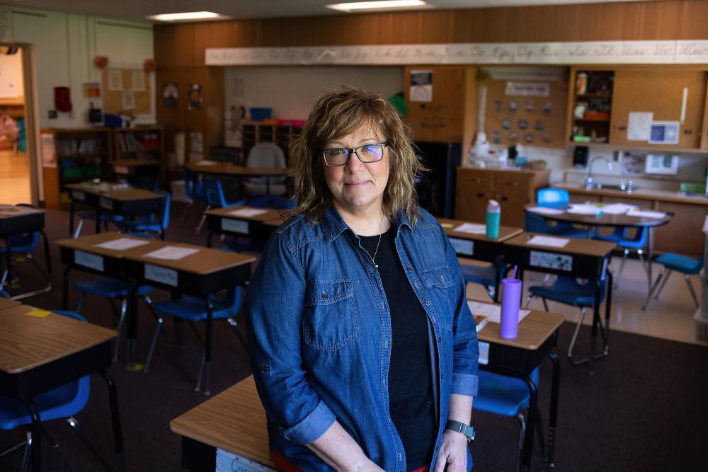 A woman wearing glasses and a denim shirt stands in a classroom. Desks and chairs are arranged around her.