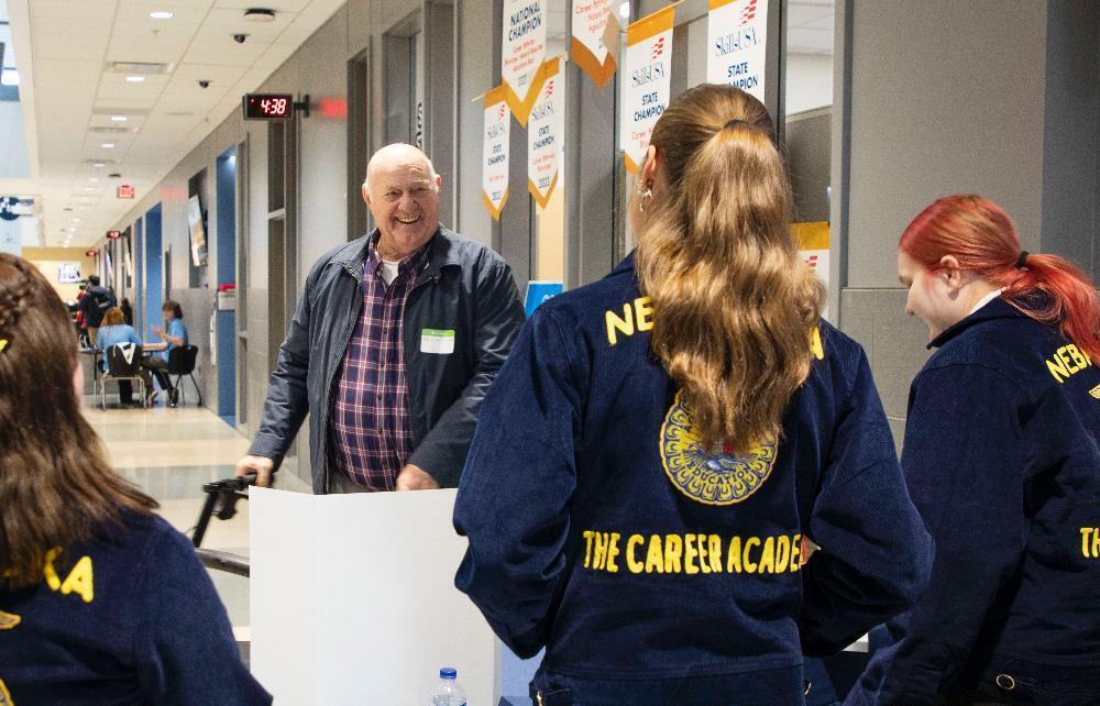 Three students and one older man with a walker are standing in the main commons area of The Career Academy's main campus. The man is wearing a light jacket and is holding onto the walker while smiling. The three students are wearing Future Farmers of America jackets and have their backs to the camera.