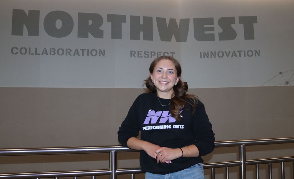 Northwest senior Amelia stands in front of a wall that has the words 'Northwest: Collaboration, Respect, Innovation' on it. She is wearing a shirt that says Northwest Performing Arts.