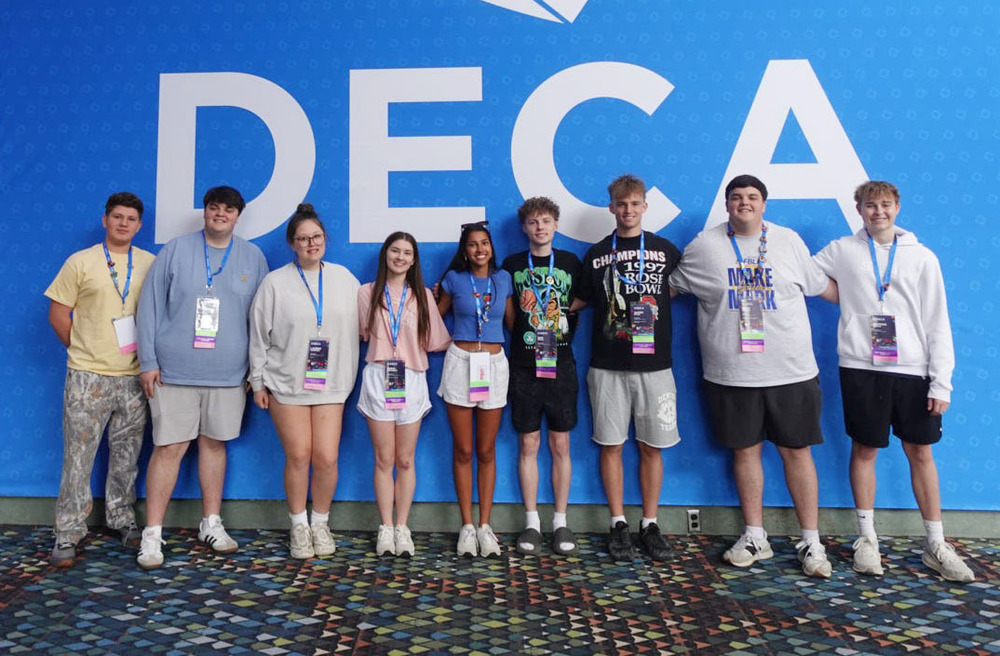 Nine Lincoln Public Schools students stand next to each other in a row at the DECA International Career Development Conference. The three girls and six boys are standing in front of a large wall that has the words DECA on it. All nine students are wearing lanyards with conference badges in them.