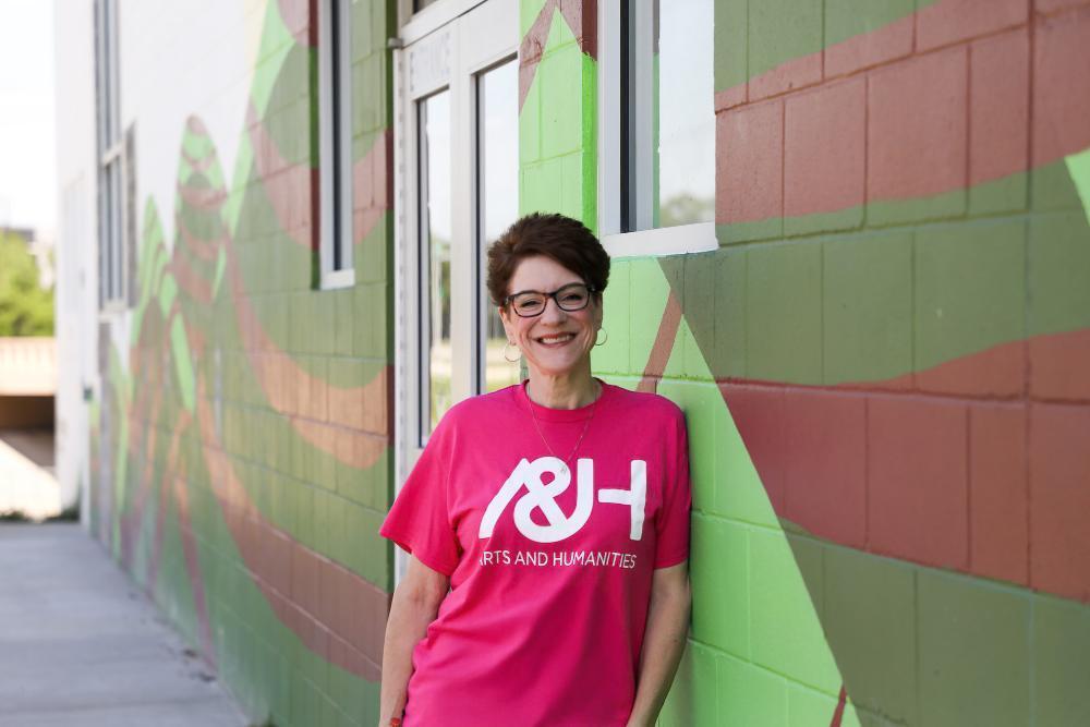 A woman, wearing a pink "A&H Arts and Humanities" shirt, leans against a colorful brick wall outdoors.
