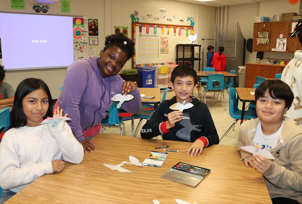 Four Elliott Elementary School student display paper birds they have made in their art class. Two boys and one girl are sitting at a table and a second girl is standing by the table in the classroom.