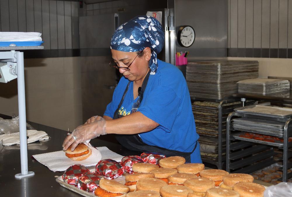 A woman in a blue uniform, apron, and patterned headscarf, wearing gloves, is assembling sandwiches in a kitchen. She places a bun on a breaded patty. A tray of similar sandwiches, some foil-wrapped, sits nearby on the stainless steel counter.