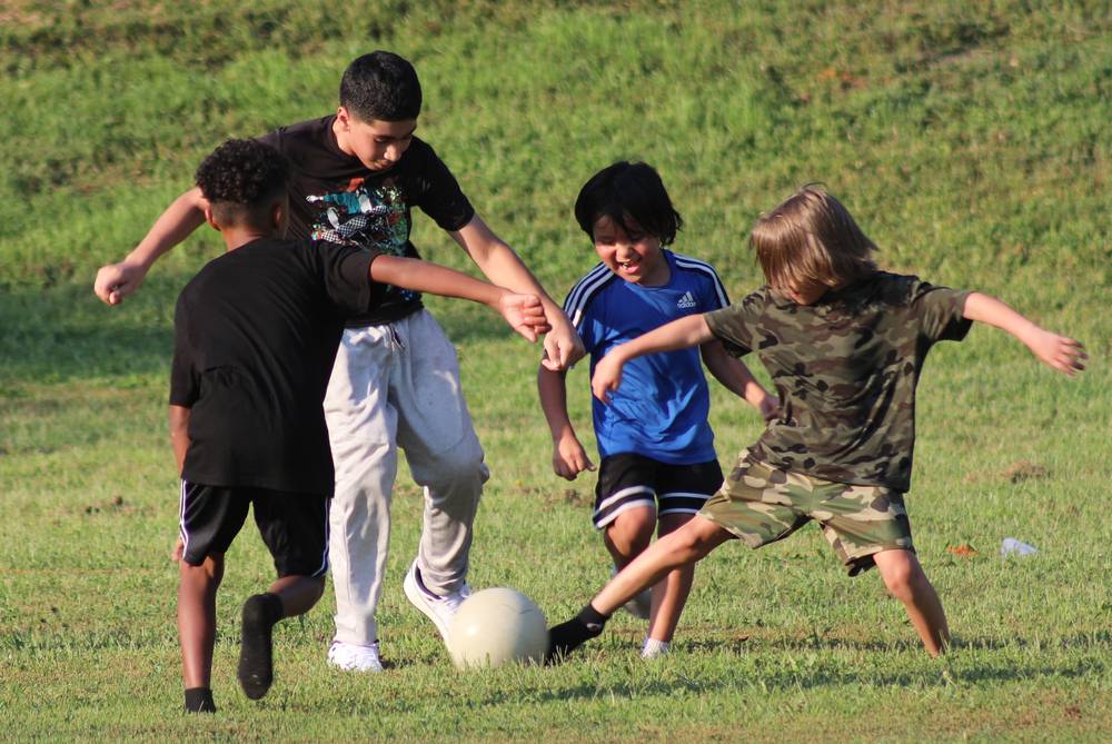 Four elementary students are attempting to kick a white soccer ball on a green field.