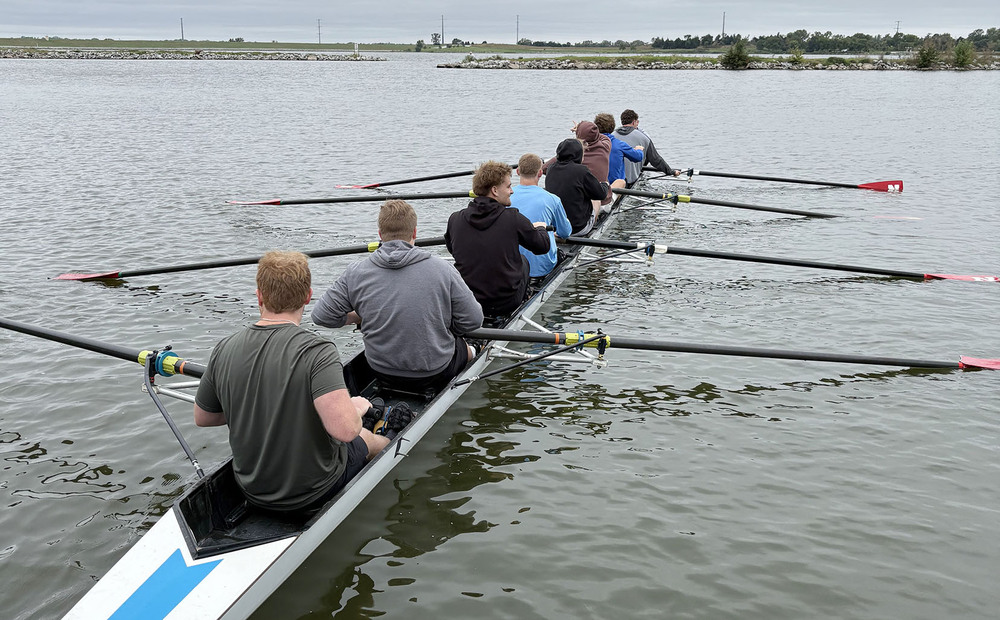 Eight East High School students are inside a long rowing boat on the waters of Pawnee Lake. They are in a row and are moving large oars together. The boat is moving away from the camera angle.